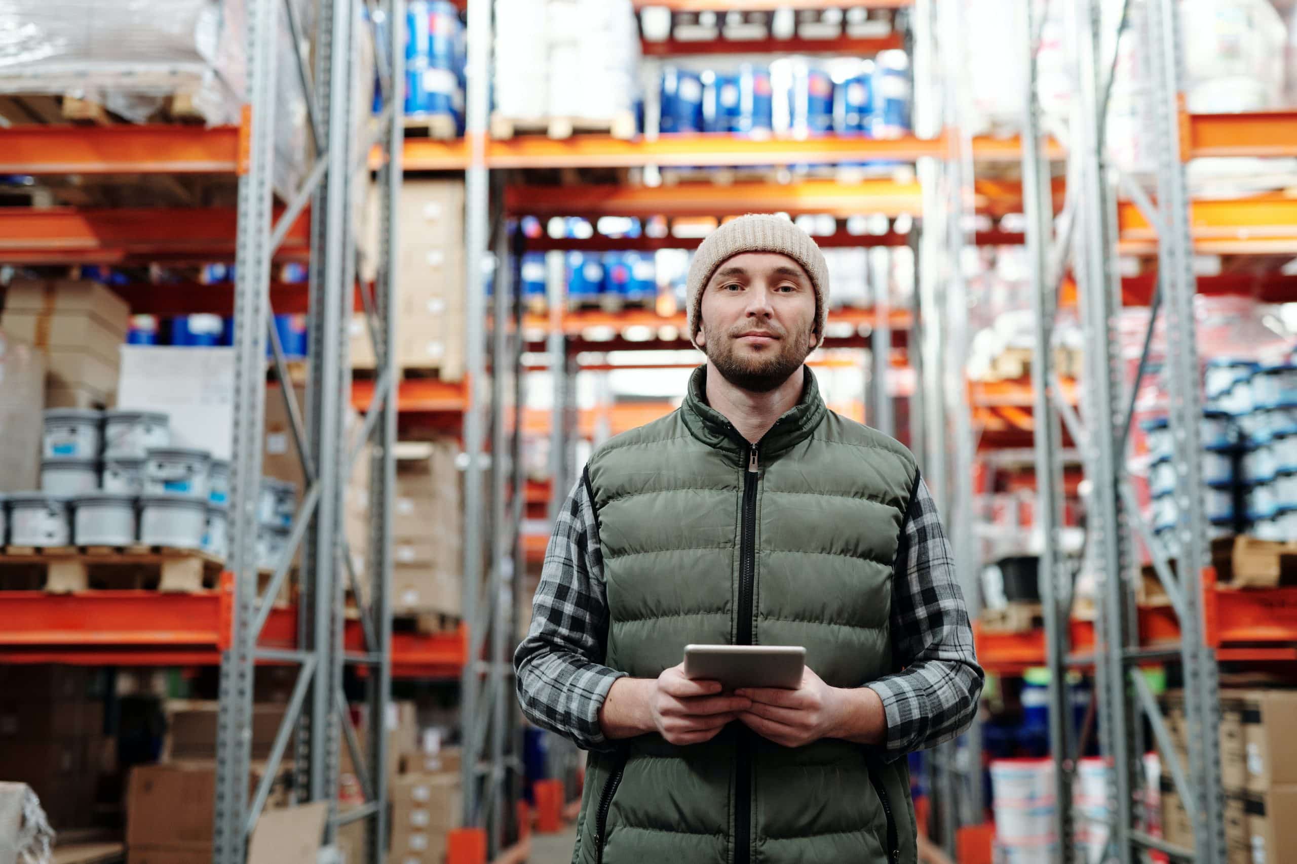 A man in a beanie and vest holds a tablet and smiles at the camera, standing in a warehouse with shelves stocked with goods, possibly engaged in a temperature mapping process.